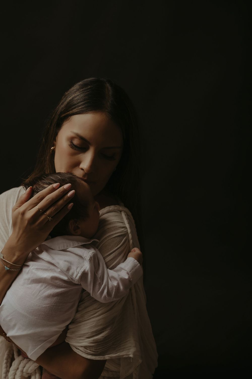 A mother tenderly kisses her newborn baby while cradling them against a dark background.