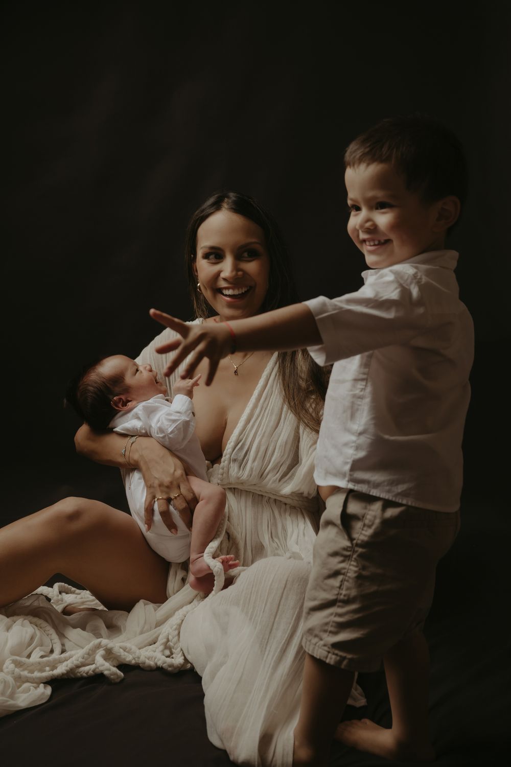 A smiling mother in a white dress holds her newborn baby while her young son reaches over to touch the infant.