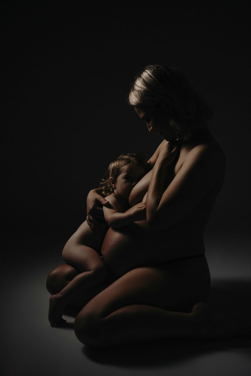 A mother breastfeeding her young child in a dark, artistic studio portrait with dramatic lighting.