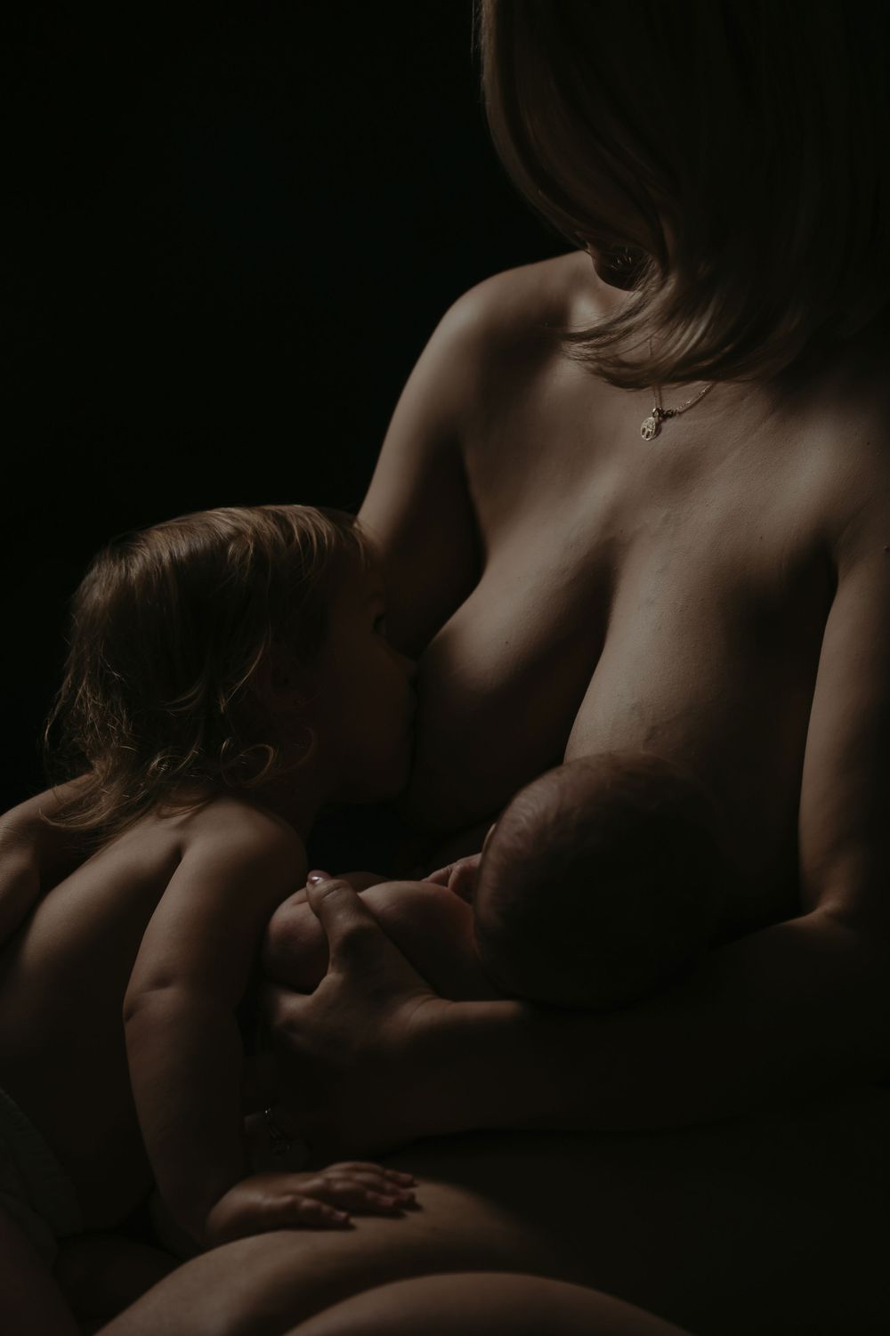 Mother breastfeeding two young children in a dark, artistic low-key portrait photograph.