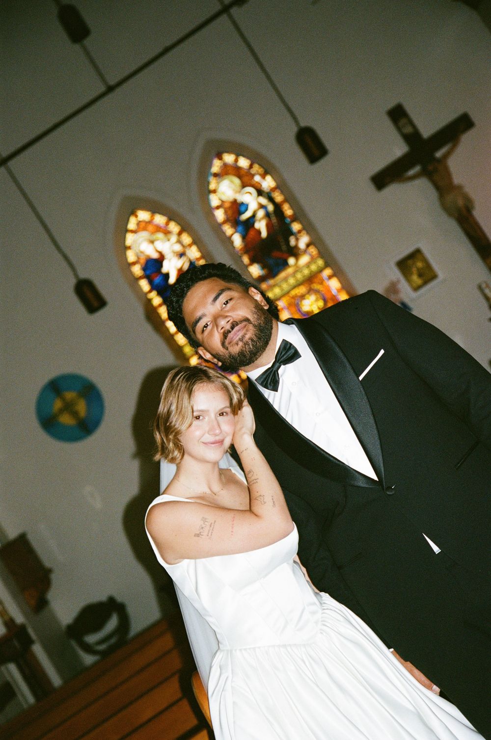 A couple in wedding attire dance and smile inside a church with stained glass windows and a crucifix visible in the background.
