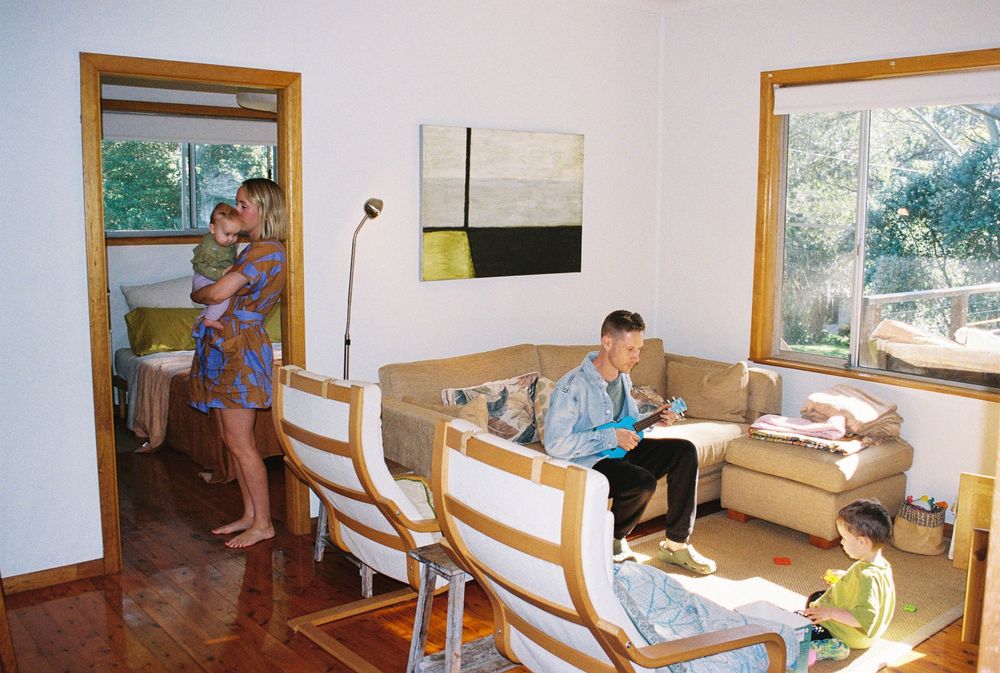 People relax in a bright living room with wooden floors, white walls, and large windows showing green foliage outside.