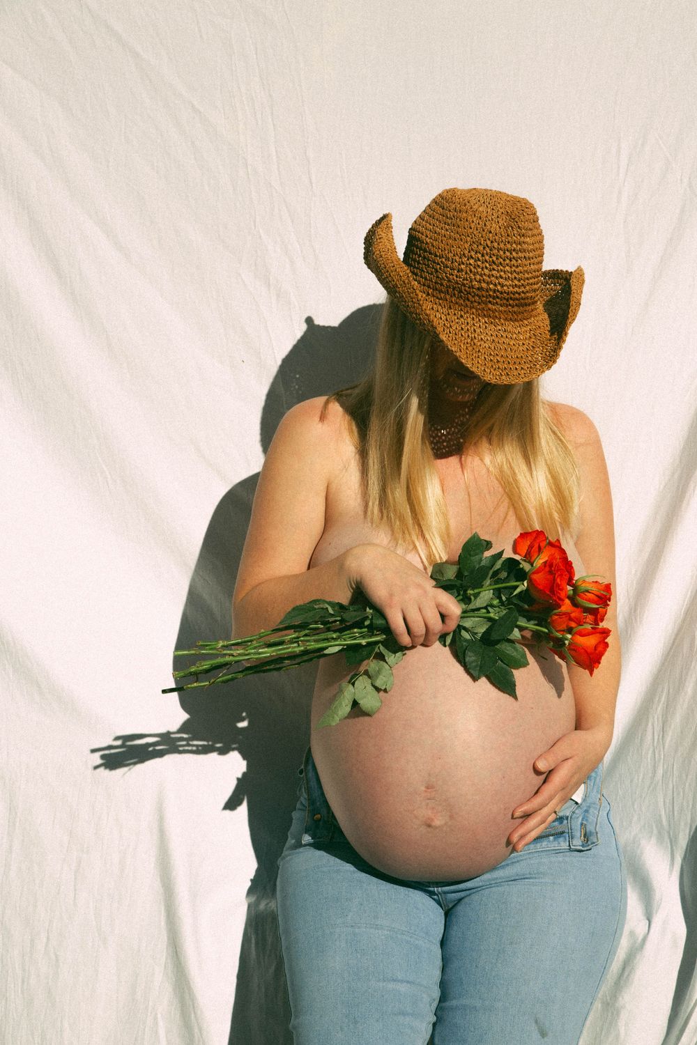 A pregnant woman in jeans and straw hat holds red roses against a white background in an artistic maternity photo.