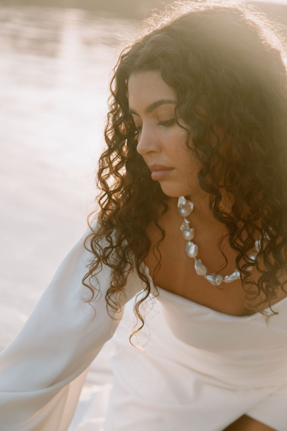A woman with curly dark hair and pearl necklace poses against a bright white background in an ethereal photoshoot.