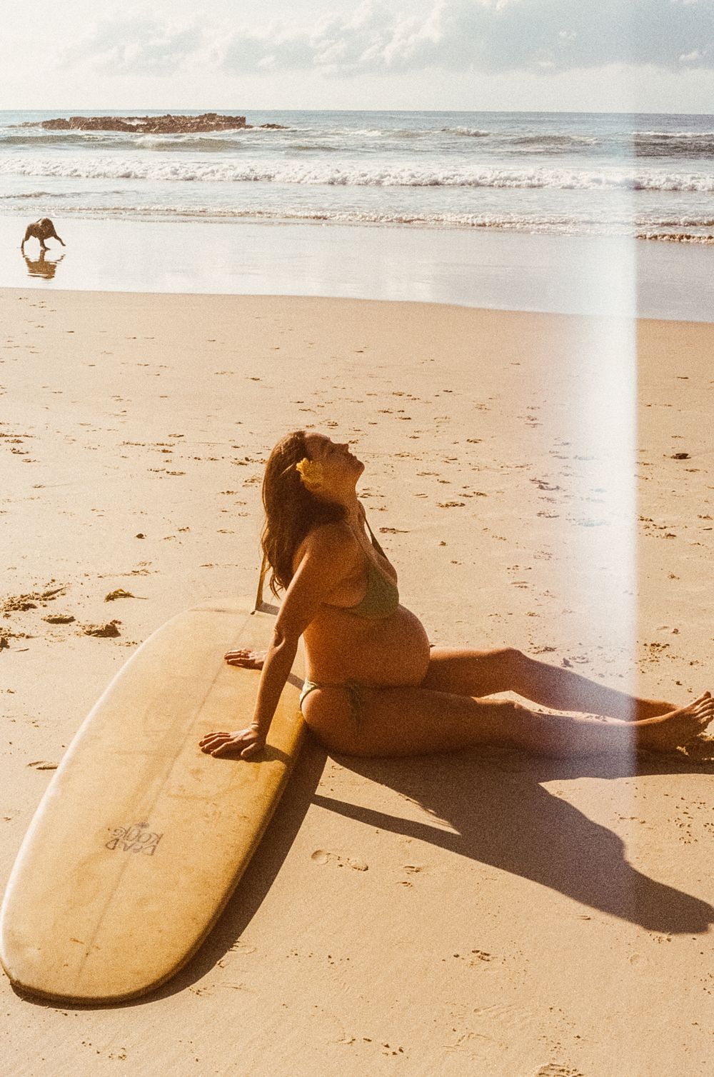 Vintage style photo of a surfer relaxing on sandy beach next to longboard surfboard with ocean waves in background.