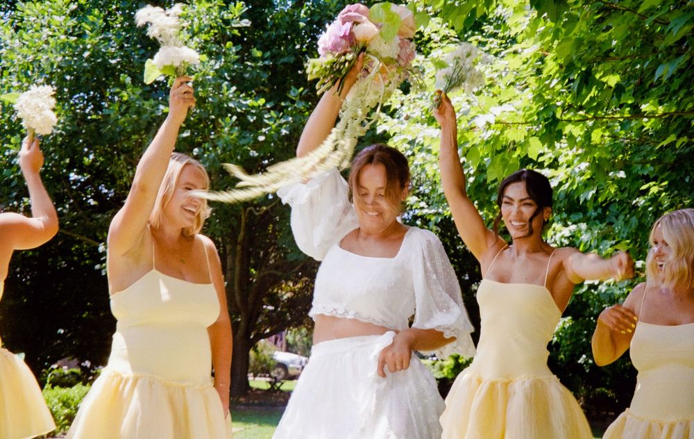 A group of bridesmaids in white and yellow dresses dance joyfully together outdoors on a sunny summer day.