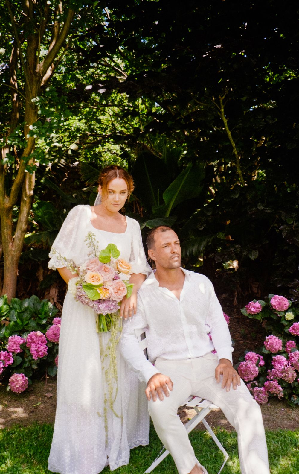 A couple in white wedding attire poses together in a garden with pink flowers and lush greenery.