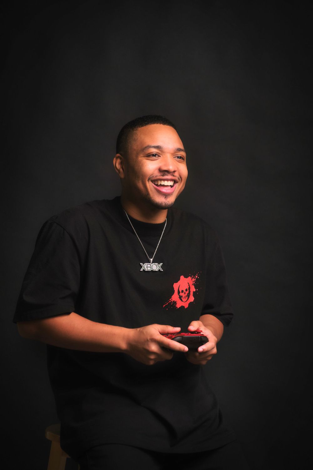 A person in a black t-shirt with a red design smiles while wearing a necklace against a dark background.