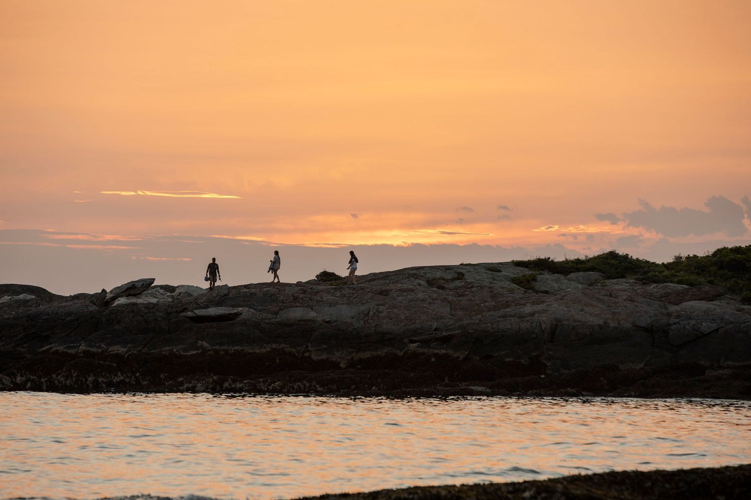Silhouettes of three people walking on rocky shoreline during a stunning golden orange sunset over calm water.