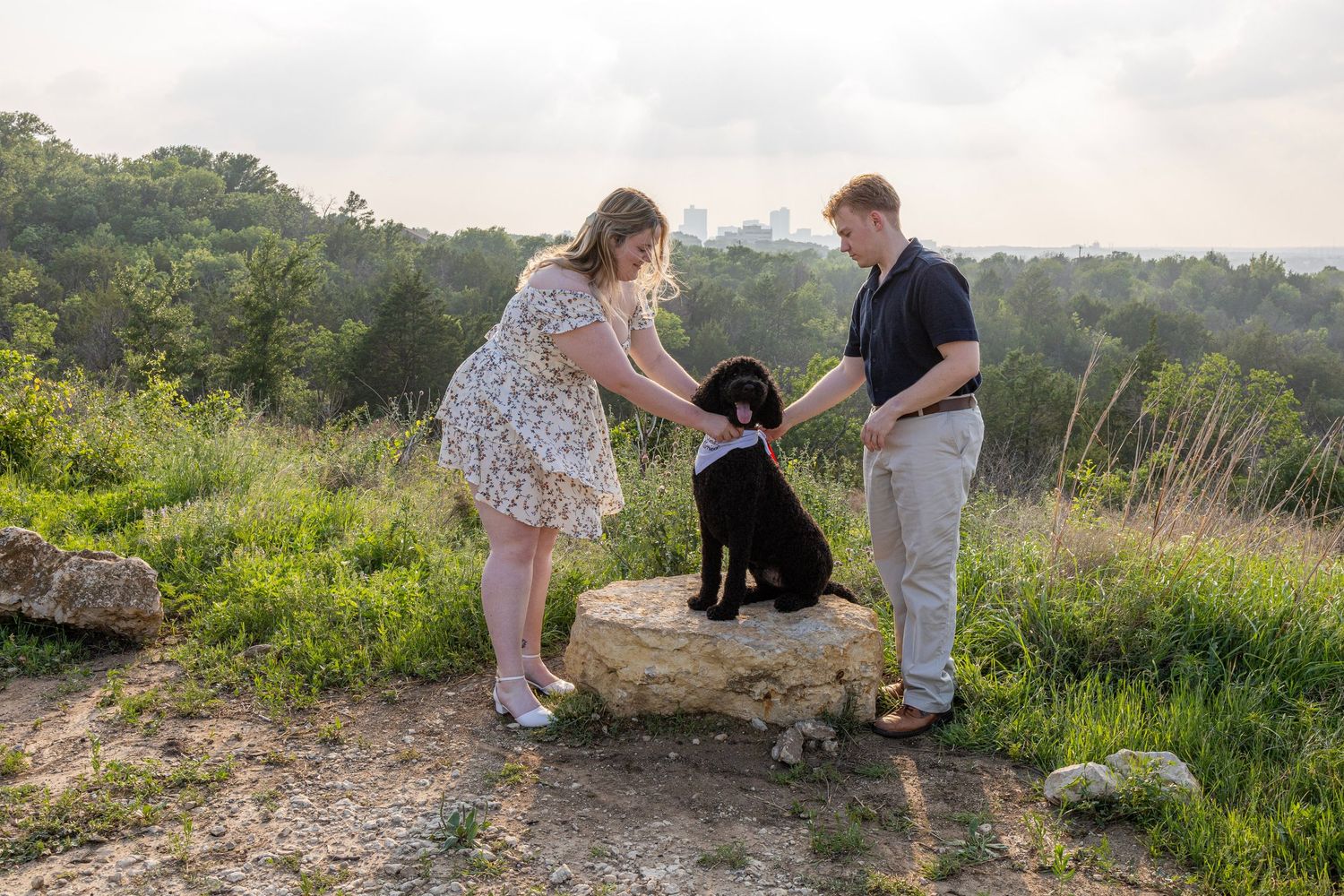 Couple petting a black dog on a rock with lush greenery and city skyline in the background.
