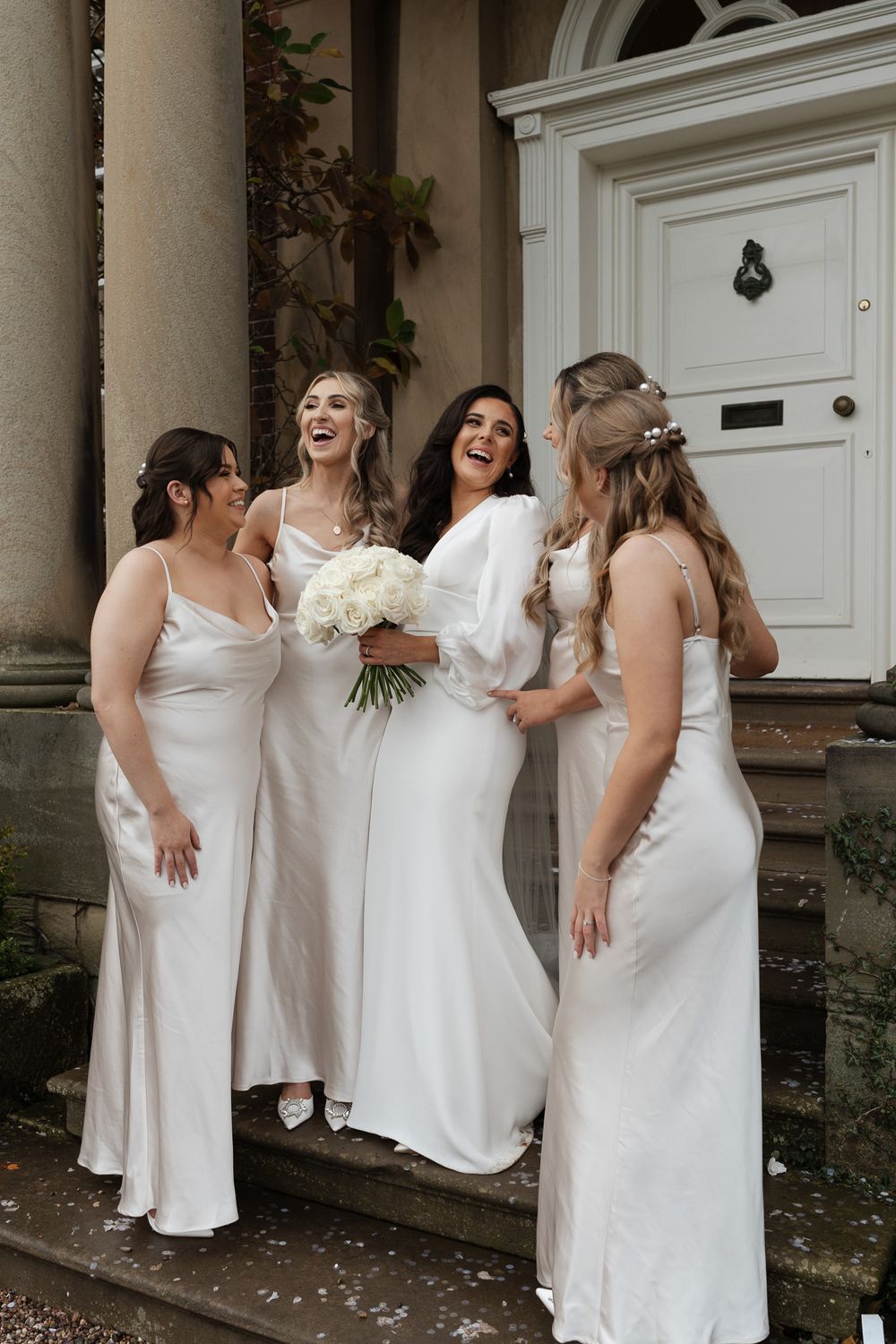 Wedding party in matching white dresses laughing together on stone steps.