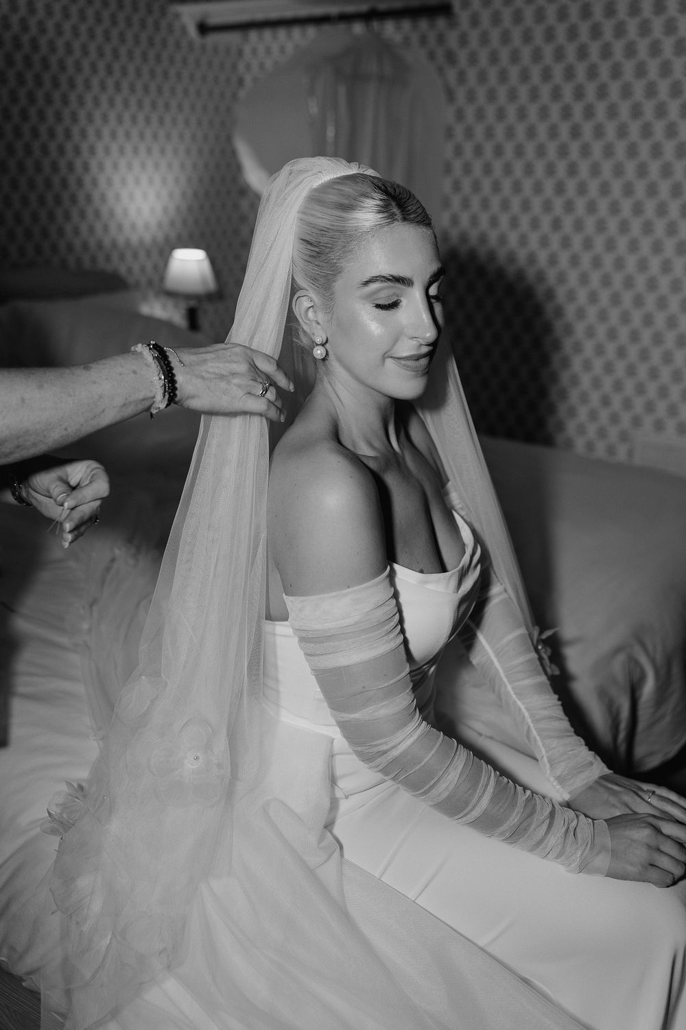 A bride in an elegant off-shoulder gown has her veil adjusted in a black and white portrait.