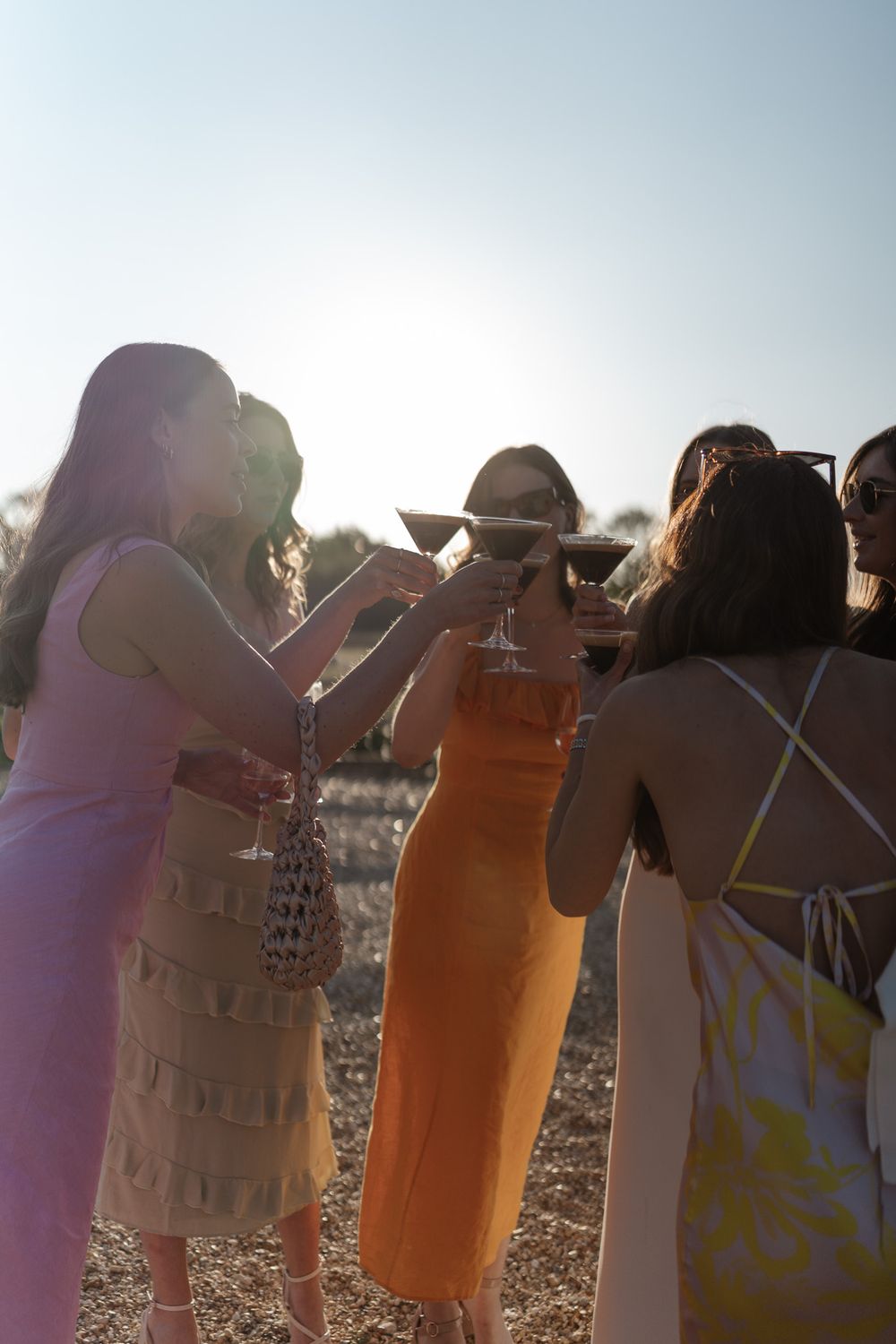 Group of women in colorful dresses toasting at sunset outdoor event.