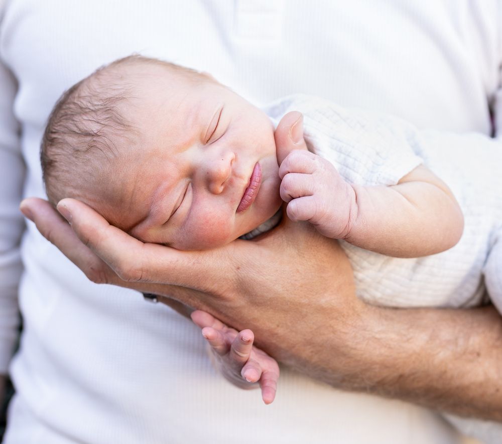 Peaceful newborn baby sleeping in father's supportive hands, wearing a white onesie.