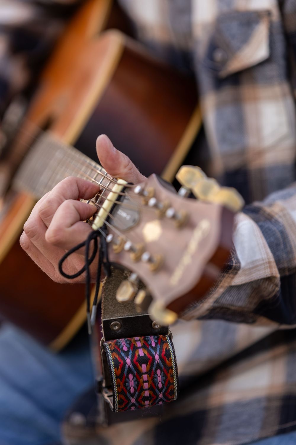 Close-up of hands playing acoustic guitar with colorful strap and plaid flannel shirt.