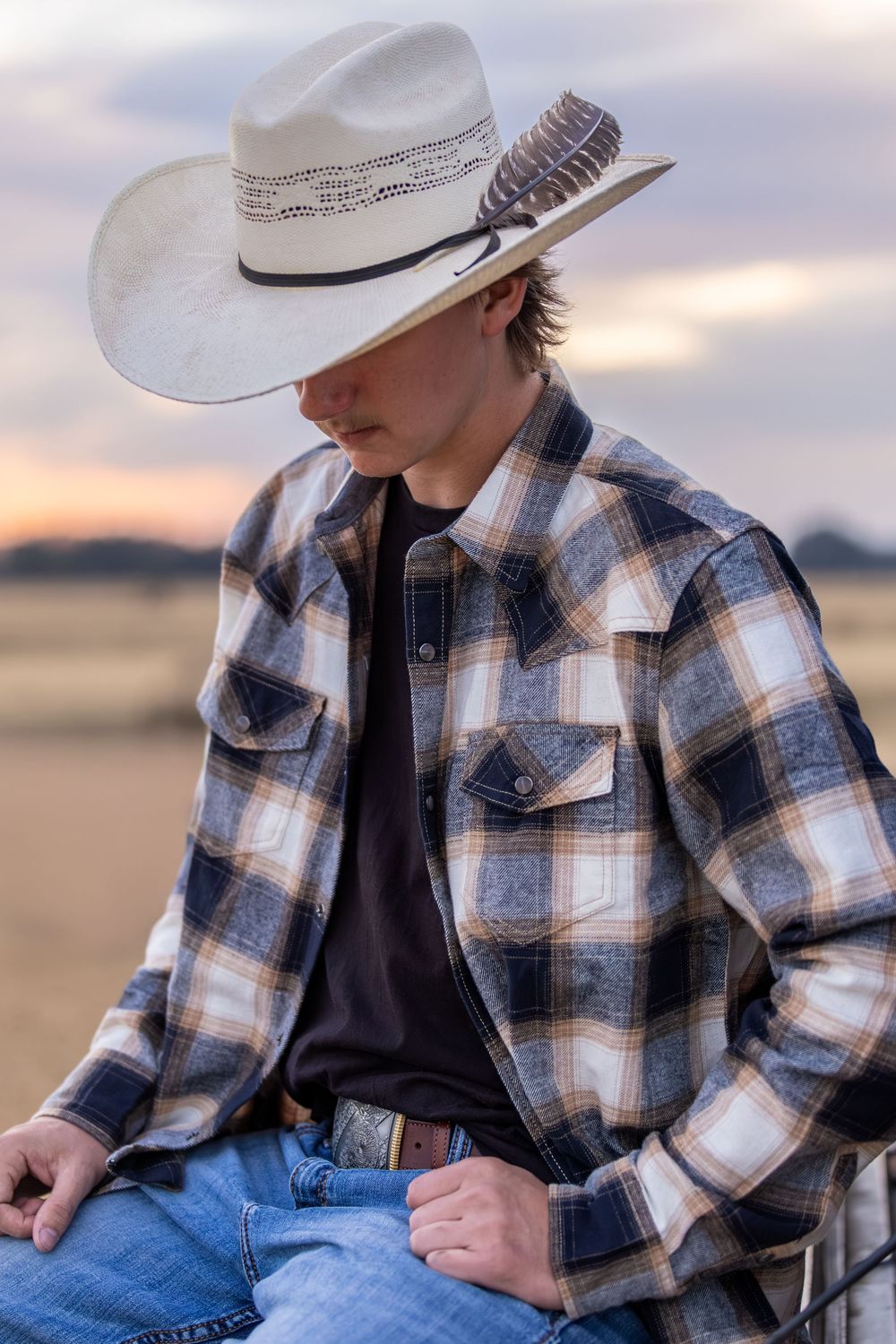 Cowboy in plaid flannel shirt and white straw hat sitting outdoors at sunset.