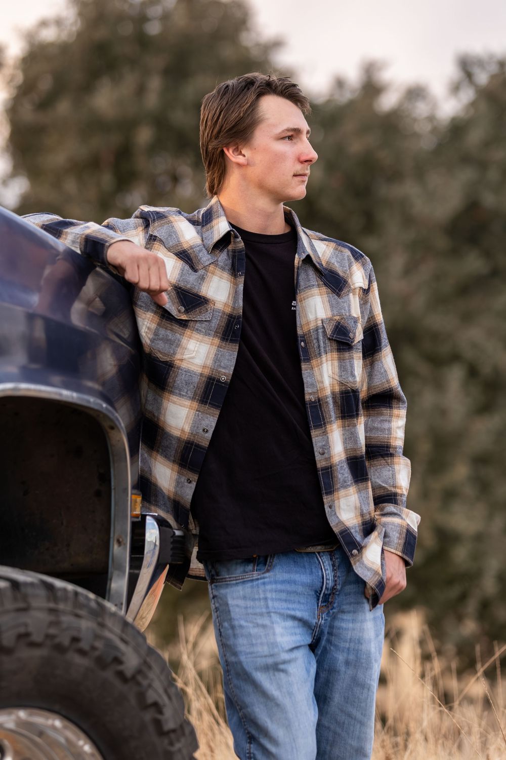 Young man in plaid flannel shirt and jeans leaning against a truck in a rural field.
