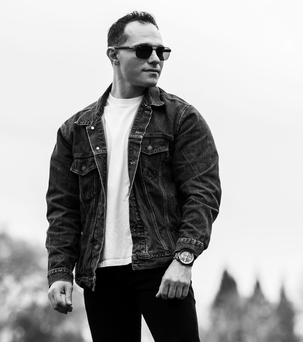 Man in denim jacket and sunglasses posing outdoors in black and white street style fashion photo.