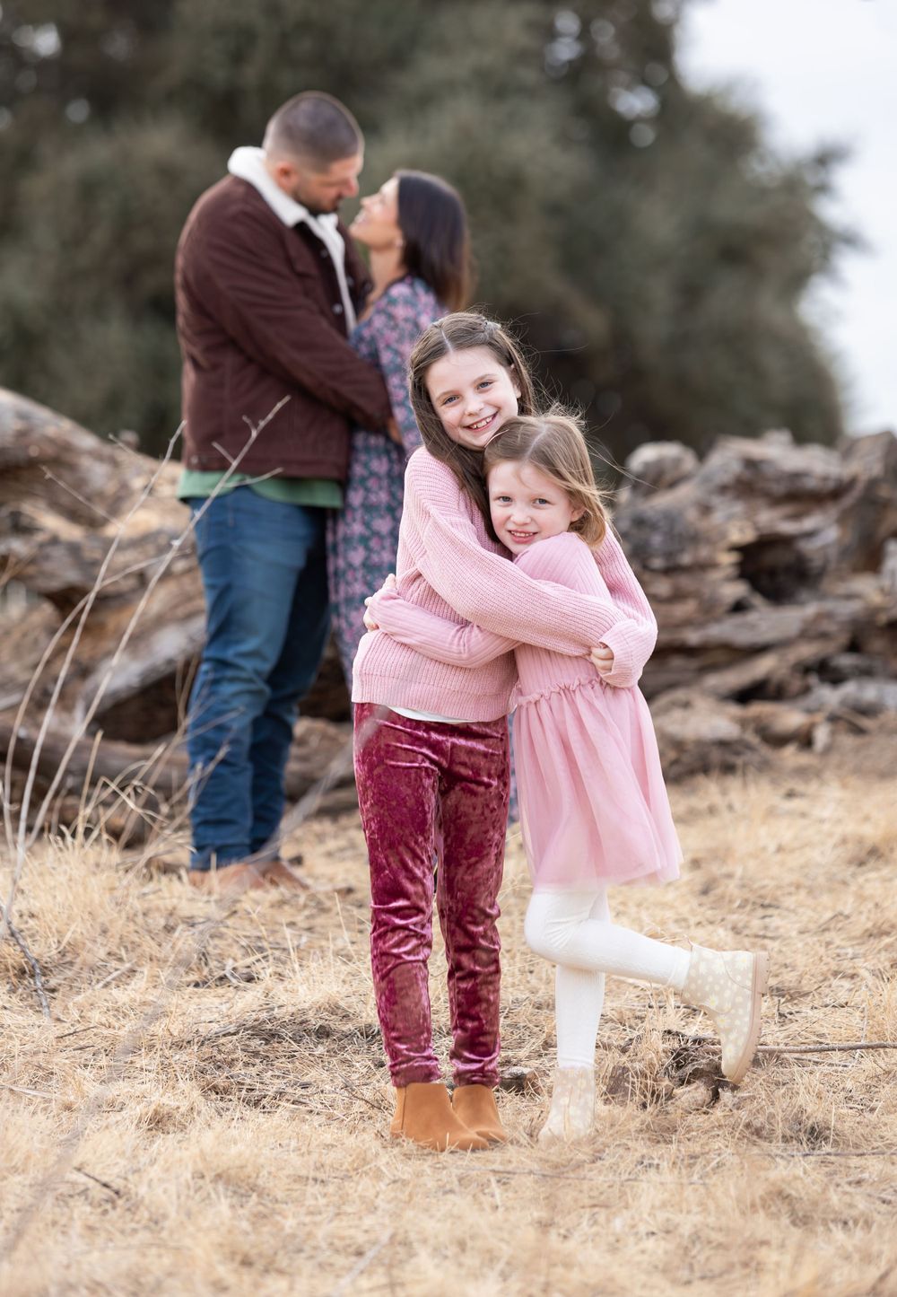 Two sisters hugging in pink outfits while parents share a kiss in the background outdoors.