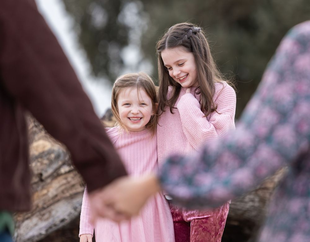 Two young girls in pink dresses laughing and hugging joyfully during an outdoor family photo session.