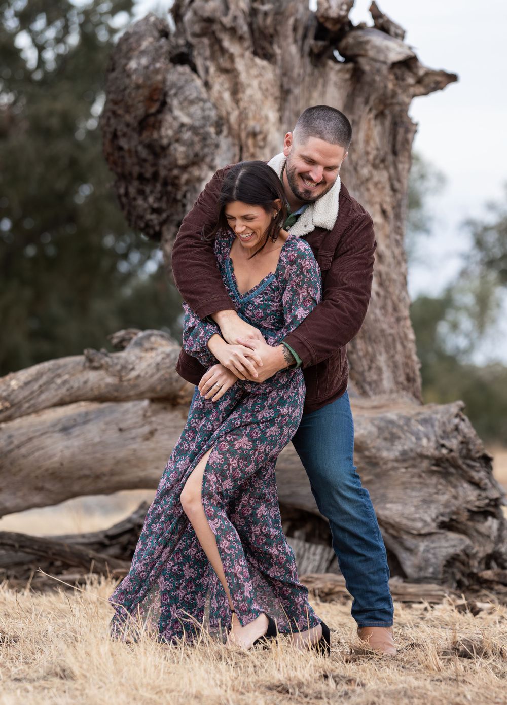 Happy couple laughing and embracing in front of a large ancient tree in a golden field.