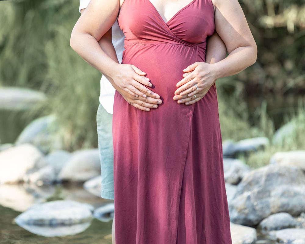 Pregnant woman in burgundy maxi dress with partner embracing her baby bump outdoors by a creek.