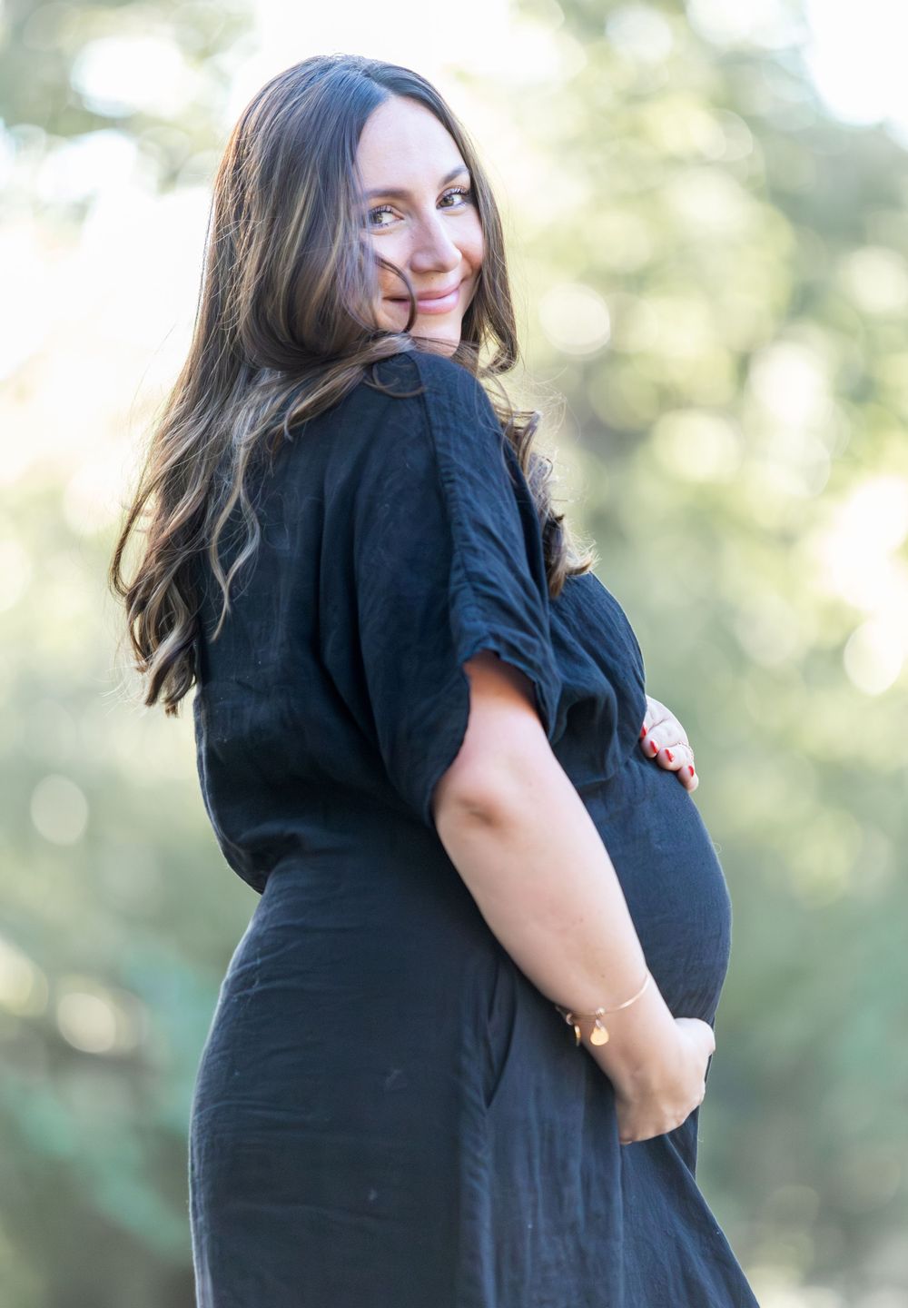 Pregnant woman in black dress holding baby bump outdoors during maternity photo session.