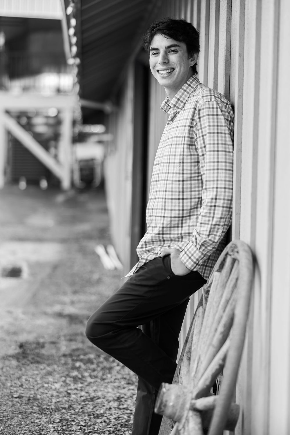 Smiling teenage boy in plaid shirt leaning against barn wall in black and white photo.