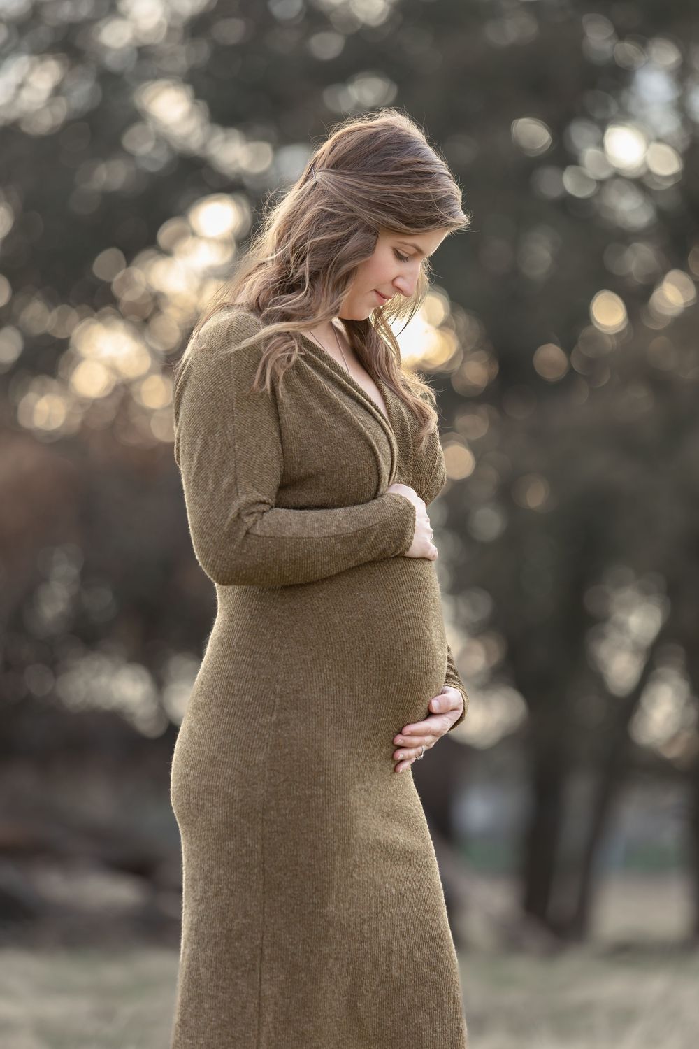 A pregnant woman in an olive green dress cradles her baby bump outdoors in soft bokeh light.
