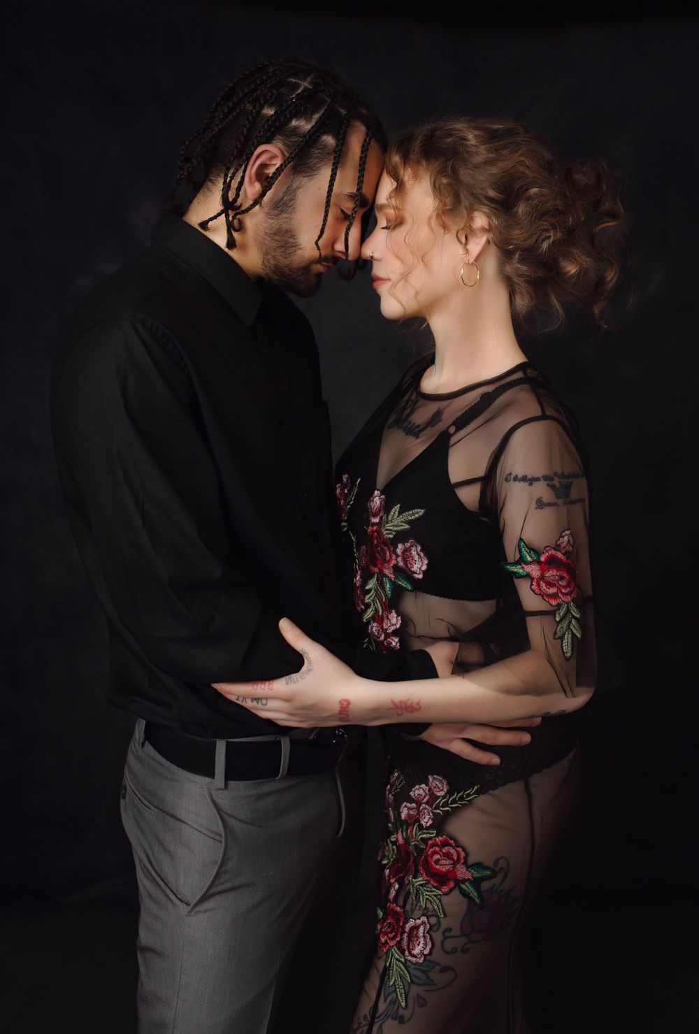 A romantic couple embraces in dim lighting against a dark background, with the woman wearing a sheer floral dress.