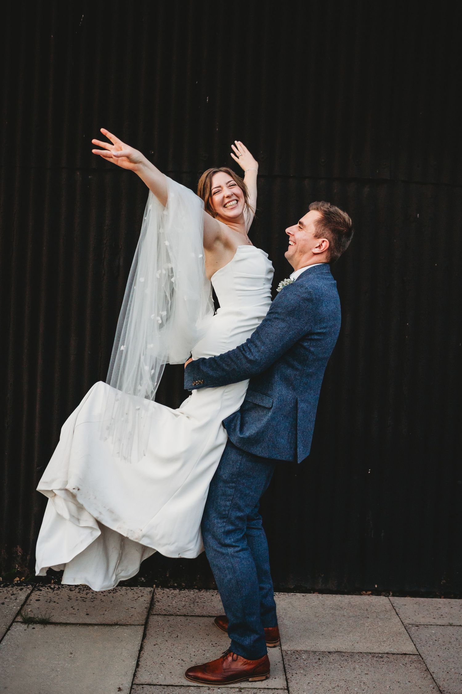Dynamic wedding couple performing playful poses against a dark wall.