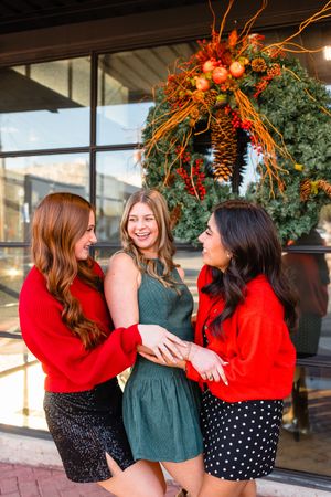 Three young women laughing together outdoors in front of a festive fall holiday wreath decoration.