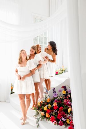 Four young women in white dresses laughing on a floral-decorated staircase in a bright white room.
