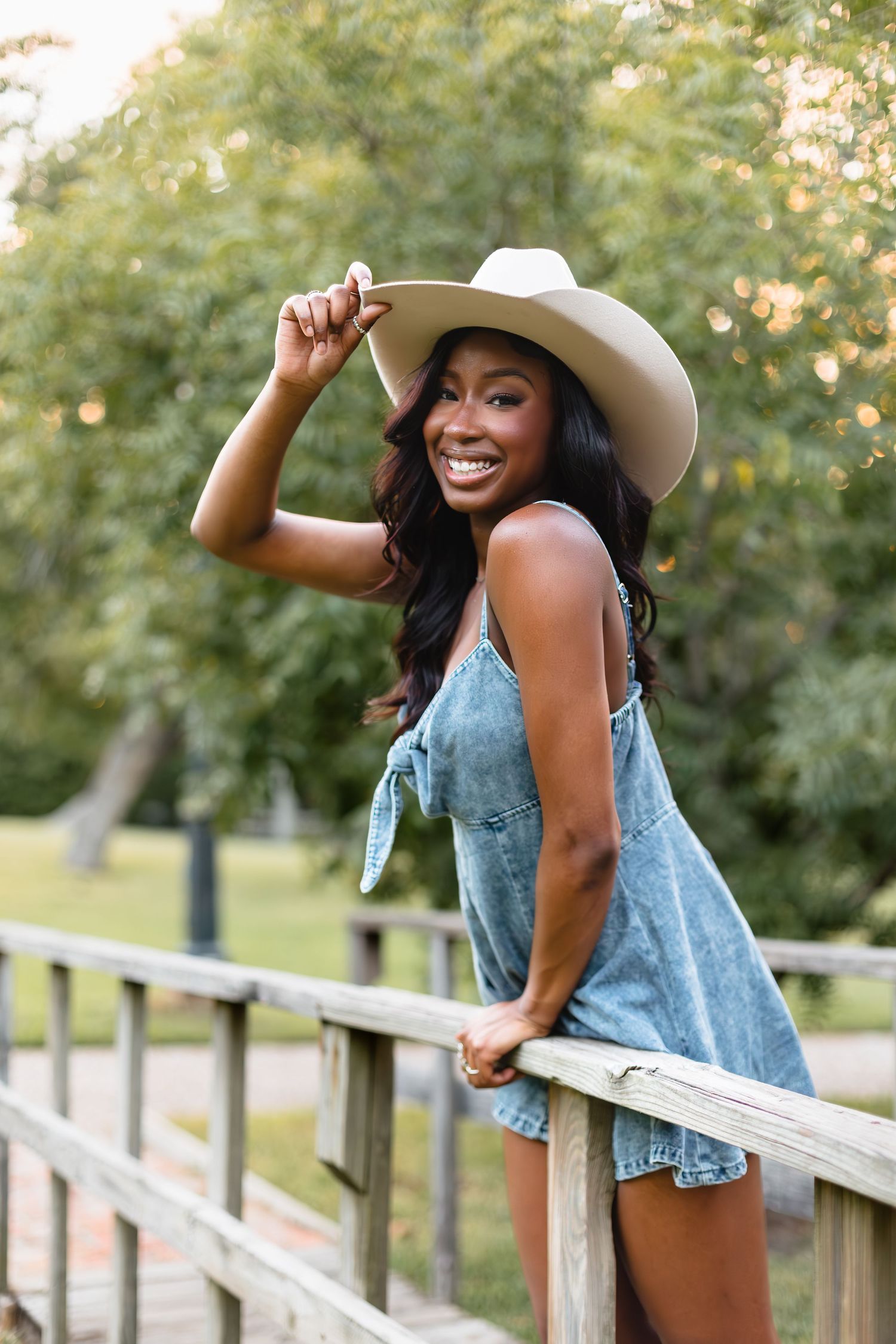 Smiling young Black woman in denim romper tips cowboy hat while leaning on wooden bridge railing.