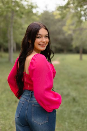 Smiling brunette teen in a hot pink off-shoulder top and jeans posing in a lush green park.