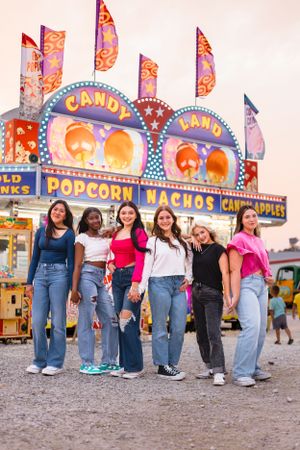 Six teenage girls posing together in front of a colorful carnival food stand at dusk.