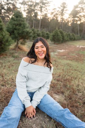 Smiling young Hispanic woman in an off-shoulder gray sweater and jeans sitting at a Christmas tree farm.