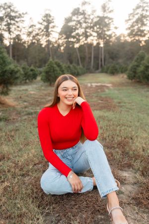 Cheerful young woman in a red sweater and jeans posing at a pine Christmas tree farm at sunset.