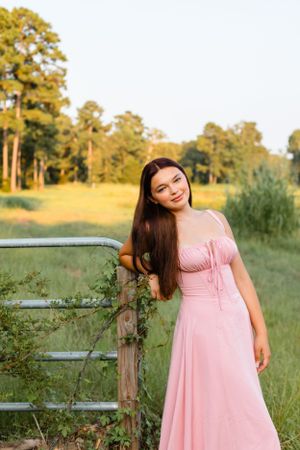 Young woman in a pink dress leaning on a rustic gate in a sunny green field at golden hour.