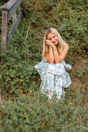 Smiling blonde girl in a blue floral dress sitting among green plants near a wooden fence outdoors.