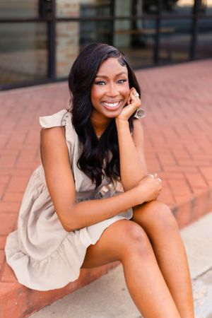 Smiling young Black woman in tan dress sits on brick steps in urban outdoor portrait.