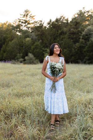 Young woman in blue floral midi dress holds white wildflower bouquet in golden hour field.