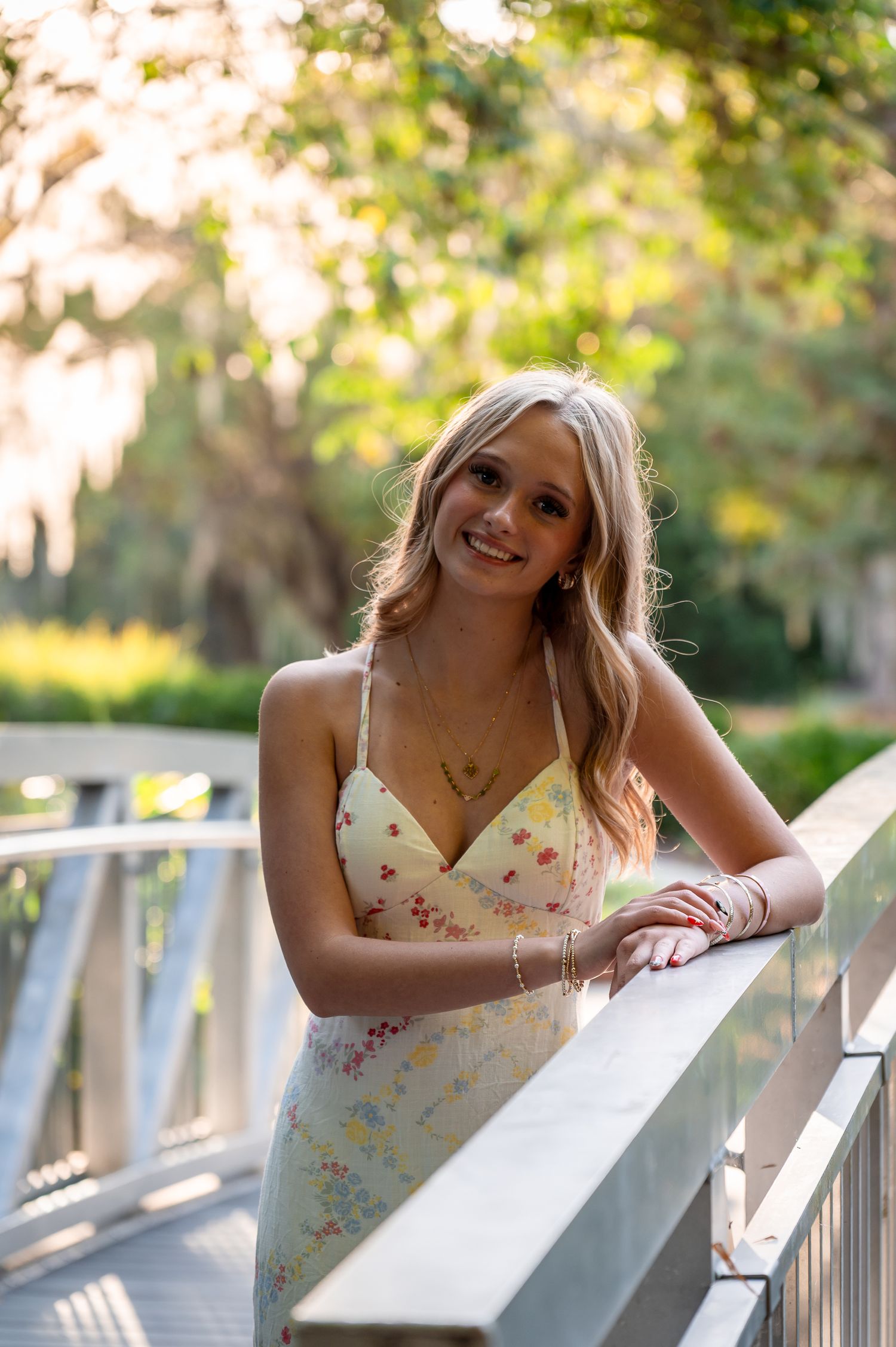 A young woman in a floral dress leans against a railing during golden hour outdoors.