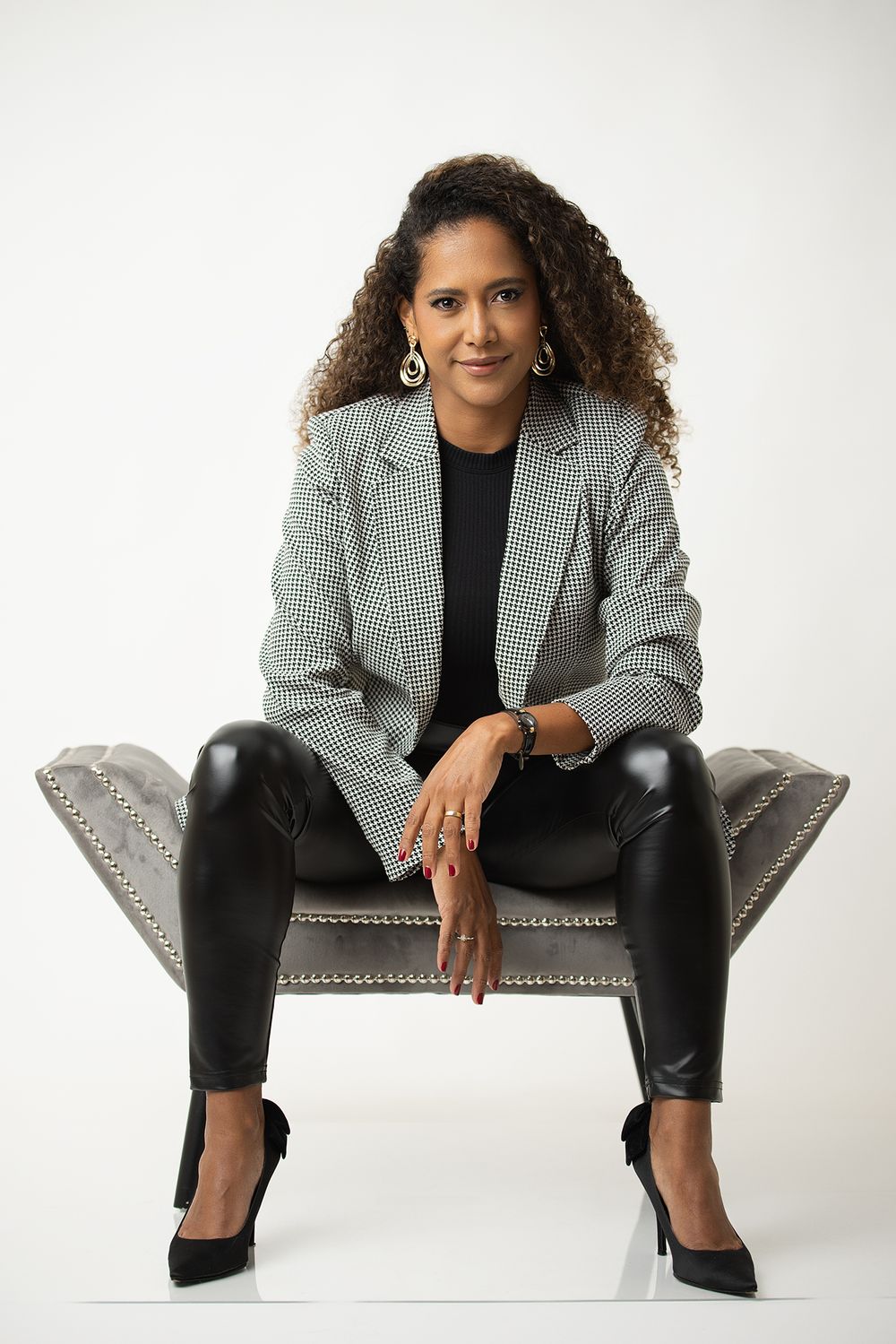 Professional portrait of someone in a houndstooth blazer and leather pants seated confidently on a gray chair. Picture by Oriana Higuera