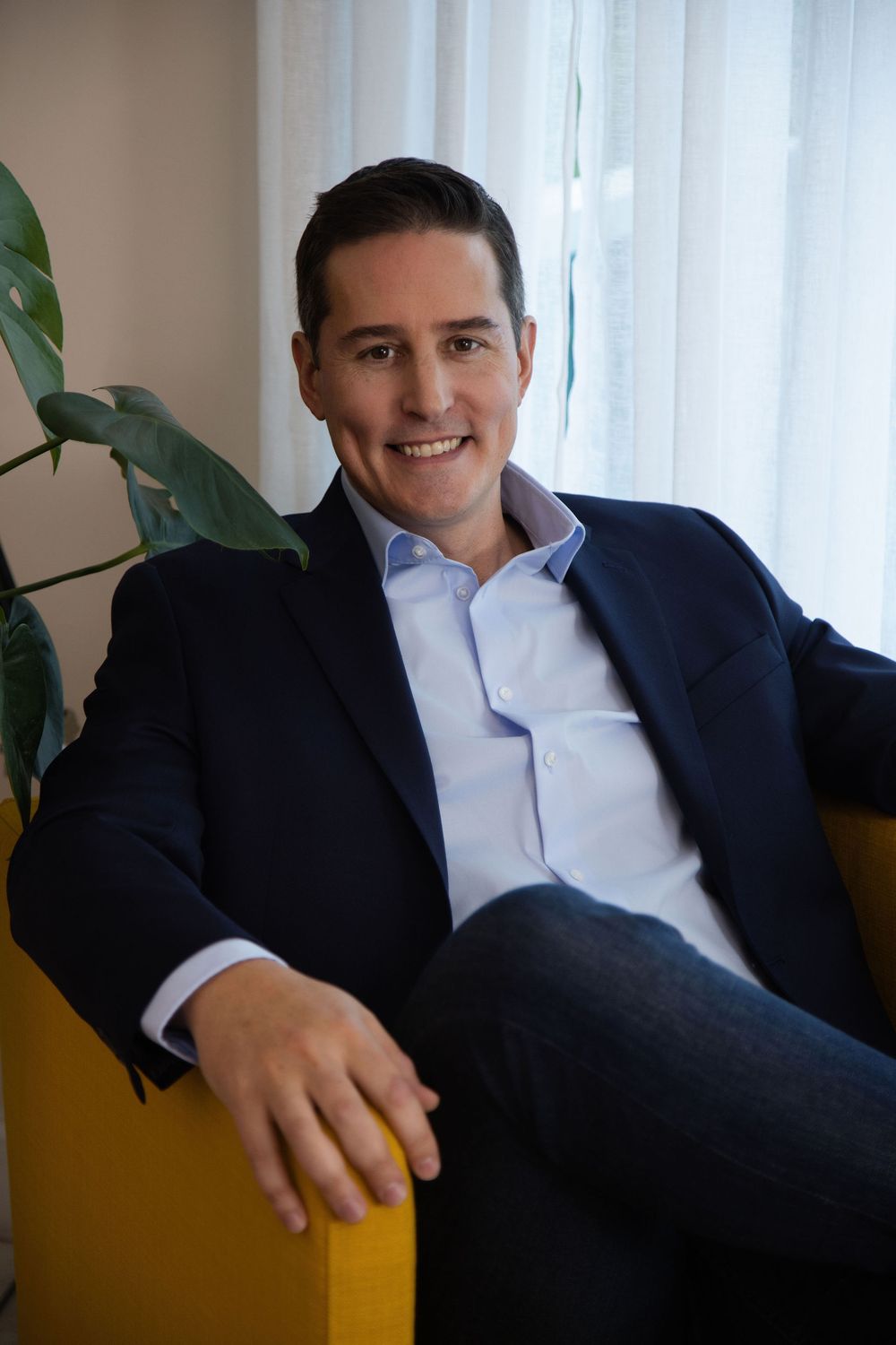 Professional portrait of someone in a navy blazer and white button-down shirt sitting casually on a yellow chair by a plant.