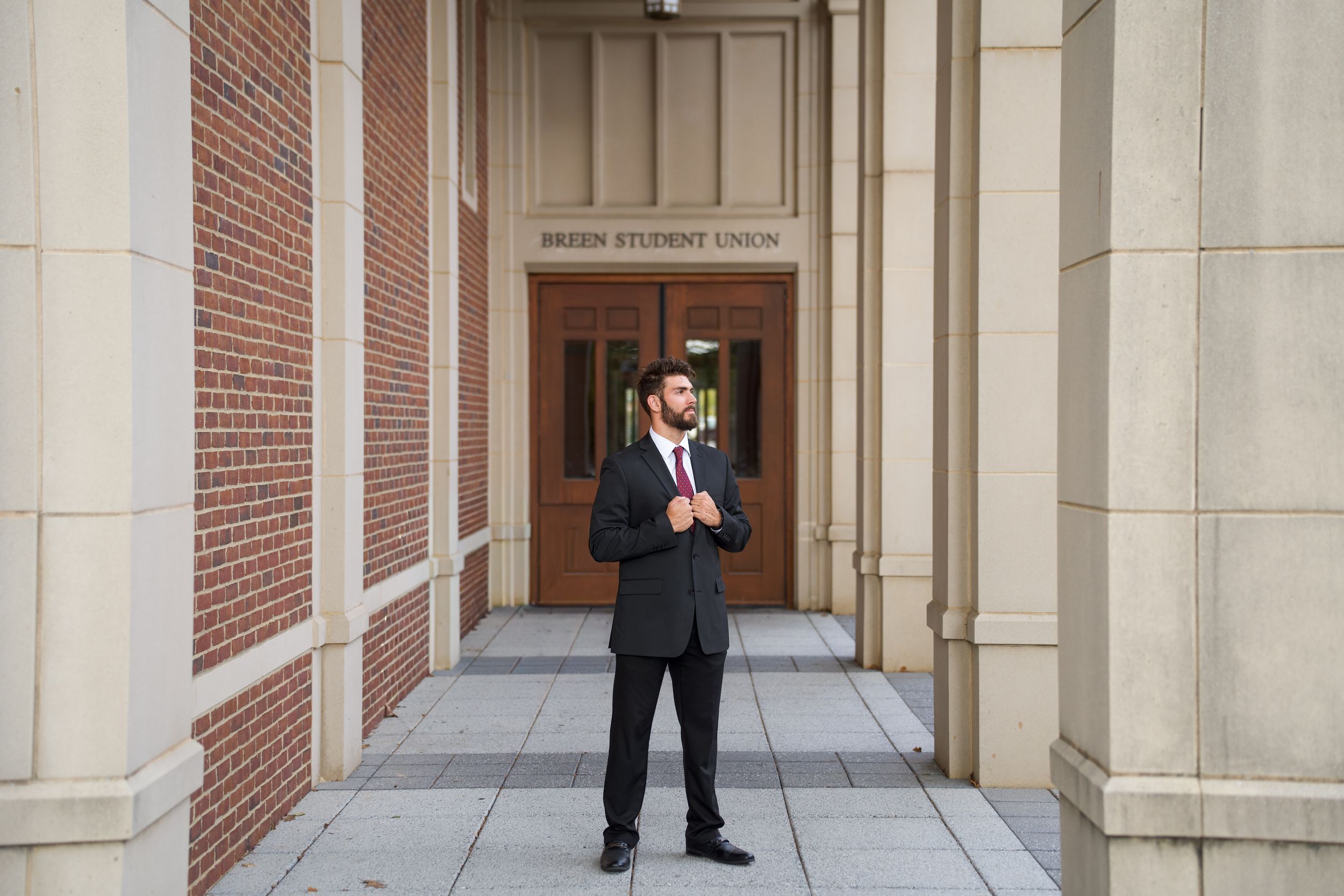 Young man in dark suit stands before brick building entrance with columns at a university student union.