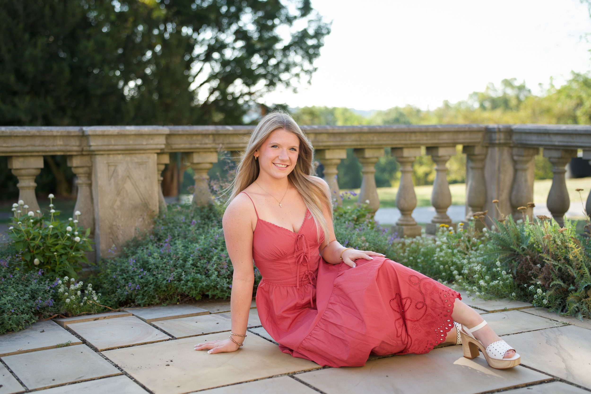 Young blonde woman in a red dress sitting gracefully on a stone patio with lush greenery and balustrade backdrop.
