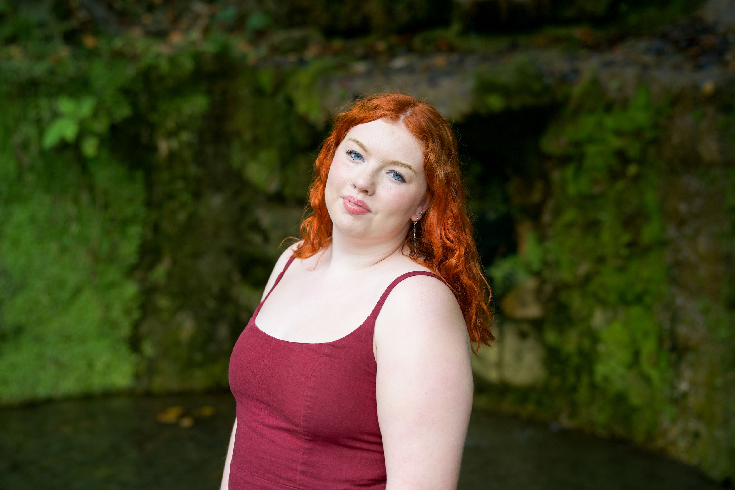 Person with red hair wearing a burgundy tank top poses outdoors against a green foliage background.
