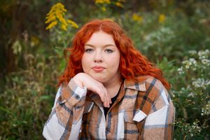 A portrait in a meadow with wildflowers showing someone wearing a plaid flannel shirt, with vibrant red hair.
