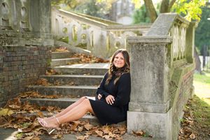 A person in a black dress sits on stone steps covered in autumn leaves near ornate architecture.